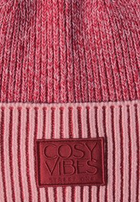 Pletená čepice v červených a růžových odstínech, s vertikálním žebrováním a texturovanou nášivkou se slovy "COSY VIBES" v tmavě červené výšivce.
