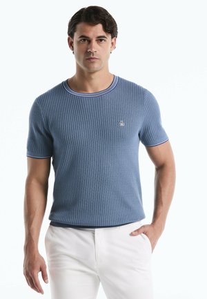 Homme portant un t-shirt bleu côtelé à manches courtes avec des bordures blanches et un pantalon blanc, debout avec une main dans la poche devant un fond blanc.