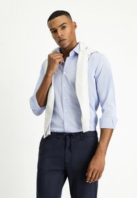 Lichtblauw overhemd met kleine patronen, opgerolde mouwen, in combinatie met donkerblauwe broek. Witte gebreide trui over de schouders gedrapeerd.