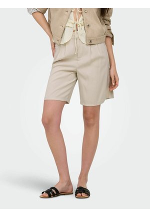 Kobieta ubrana w beżowe krótkie spodenki do kolan, krótki beżowy żakiet, kremowy krótki top i czarne płaskie sandały z odkrytymi palcami, stoi na białym tle.