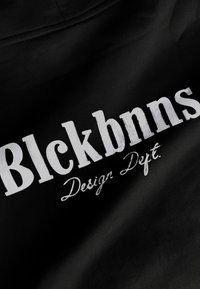 Zwarte stof met opvallende witte tekst waarop staat "Blckbnns Design Dept." De tekst heeft een verweerde uitstraling, wat de visuele textuur versterkt.