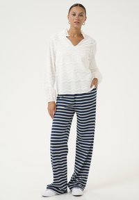Blusa bianca a maniche lunghe, con texture e vestibilità ampia, abbinata a pantaloni larghi a righe blu scuro. Le scarpe sono sneaker bianche.