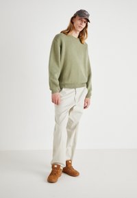 Pulover tricotat verde deschis cu margine cu nervuri, asortat cu pantaloni bej. Pantofi sport din piele întoarsă maro cu șireturi plate. Căciulă gri cu detaliu logo.