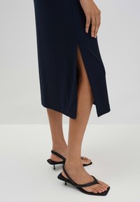 Navy-blaues, figurbetontes Kleid mit einem seitlichen Schlitz bis zum Oberschenkel; kombiniert mit schwarzen Riemchensandalen mit quadratischer Spitze und schmalem Absatz.
