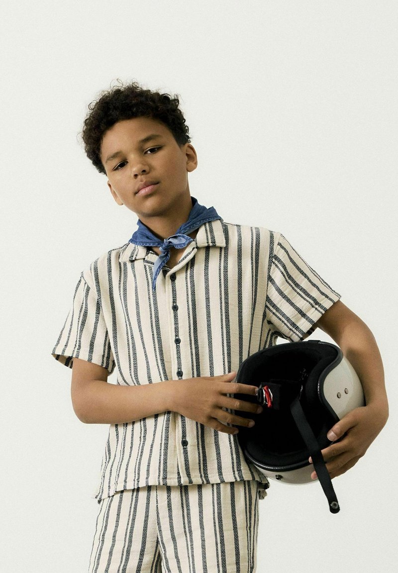Camicia a maniche corte a righe beige e blu navy, abbinata a pantaloni corti, con un casco grigio dotato di cinturino nero e imbottitura interna.