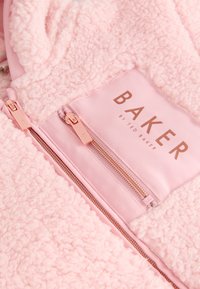 Nærbillede af blødt lyserødt fleecestof med to roseguld lynlåse og en lyserød mærkat med teksten "BAKER by Ted Baker".