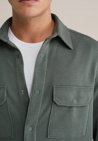 Sötétzöld gombos ing, puha anyagból készült, két elülső zsebbel, patentos gombokkal és klasszikus gallérral. Fehér póló fölé viselve.