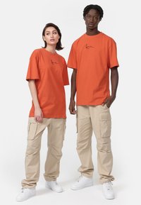 Orange bomuld t-shirts med korte ærmer og sort logo. Lysebrune cargo bukser med lommer. Hvide sneakers fuldender det afslappede look.
