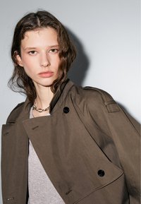Brown mid-length jacket made of smooth fabric, featuring a double-breasted design, large black buttons, and structured collar.