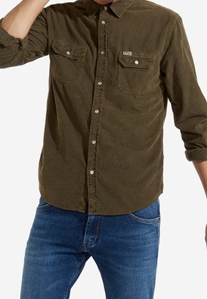 Mężczyzna w oliwkowej, długorękawowej koszuli na guziki z dwoma kieszeniami na piersi i niebieskich dżinsach.