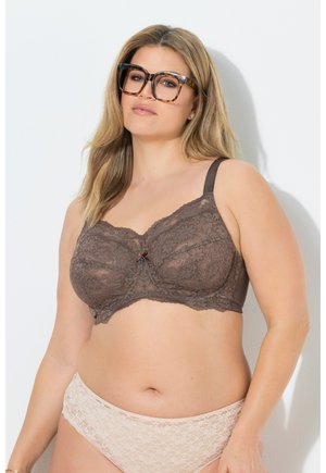 Kvinde med blondt hår iført store briller med skildpaddeskaller, en brun blonde-bh og beige blondeundertøj, stående mod en hvid væg.