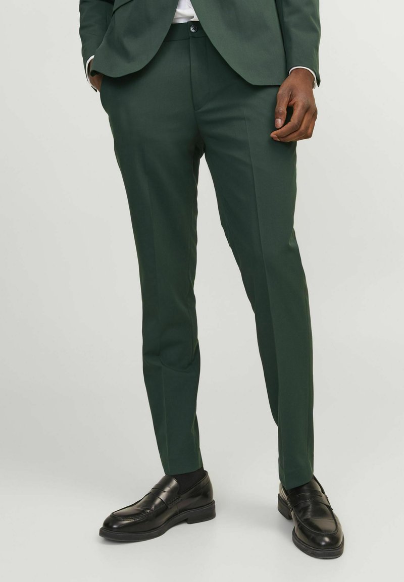 Pantalones de traje de color verde oscuro, confeccionados en tejido suave, con corte ajustado, sin pliegues en la parte frontal y con un sutil acabado. Combinados con mocasines negros.