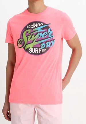 Różowa koszulka z krótkim rękawem z okrągłym wzorem z napisem "Super Dry Surf Co." w niebieskim, czarnym i zielonym kolorze z przodu.