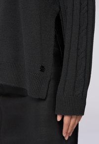 Maglione nero lavorato a maglia con un motivo a cavo testurizzato sulle maniche, dotato di un colletto e polsini a coste, abbinato a un capo nero.
