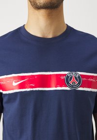 Tamsiai mėlynas marškinėliai su drąsia raudona horizontalia juostele, baltais akcentais, Nike logotipu ir Paryžiaus Šventojo Germeno emblema centre. Trumpomis rankovėmis.