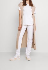 Béžový pletený cardigan s bočními kapsami a knoflíky, bílé tričko s květinovou výšivkou a bílé džíny spojené s bílými teniskami.