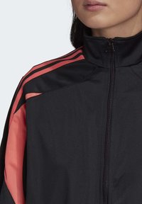 Svart zip-up jacka med korallfärga accenter och tre horisontella ränder över axlarna, tillverkad av ett smidigt och lätt material.