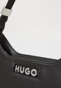 Nærbilde av svart lærveske-rem med sølvspenne og HUGO metalllogo-plakett.