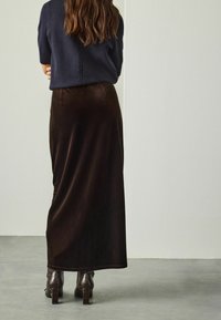 Donna in piedi con le spalle rivolte verso la fotocamera, che indossa un maglione blu scuro, una lunga gonna marrone e stivali in pelle marrone alti fino alla caviglia.