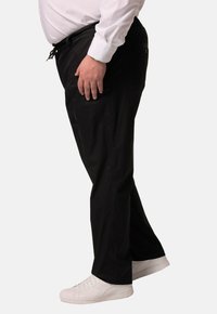Zwarte, elastische broek met zijzakken en verstelbare trekkoord, gecombineerd met witte sneakers, weergegeven in zijprofiel.