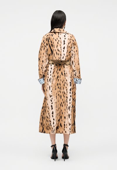 Γυναίκα με παλτό με ζώνη και animal print με βούλες, με γυρισμένα μπλε μανίκια και μαύρα παπούτσια με ψηλό τακούνι και λουράκι στον αστράγαλο, κοιτάζοντας προς τα πίσω.