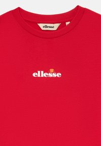 Camiseta de algodón roja con cuello redondo. Presenta un logo blanco de "ellesse" en la parte izquierda del pecho, diseño sencillo, textura suave.