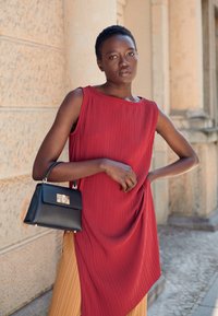 O vestido vermelho pregueado apresenta um corte solto com uma bainha assimétrica. Uma pequena bolsa preta com detalhe de fecho complementa o visual.