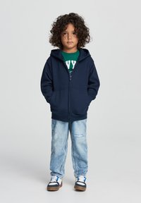 Felpa con zip blu navale e tasca frontale, indossata sopra una maglietta verde. Jeans chiari strappati e sneakers con dettagli blu navale completano l'outfit.