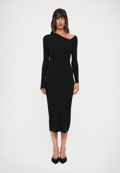 Vestido negro de punto acanalado con un hombro al descubierto y una manga larga, diseño ajustado y longitud a mitad de pantorrilla. Se lleva con tacones negros puntiagudos.