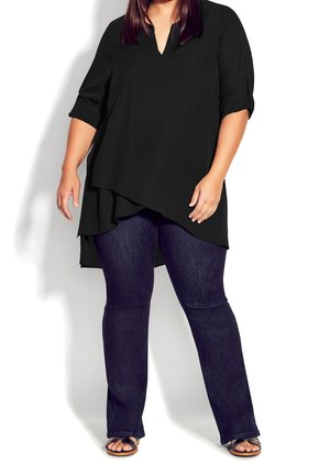 Musta tunika, jossa on v-kaula-aukko ja kolme neljäsosan hihaa, laskeutuu epäsymmetrisesti tummansinisten leveälahkeisten farkkujen ja mustien sandaalien päälle.