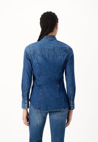 Persona con capelli intrecciati vista da dietro, che indossa una camicia di jeans blu aderente e jeans blu abbinati, su uno sfondo bianco semplice.
