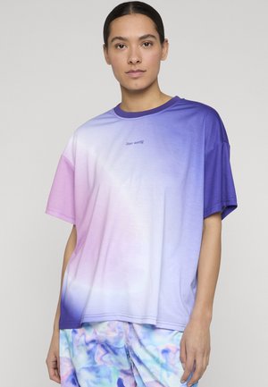 Kvinne med mørkt hår som har på seg en oversized T-skjorte med gradient fra lilla til hvit, og bukser med pastellblå, lilla og rosa abstrakt mønster.