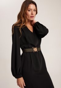 Czarna sukienka z długimi bufiastymi rękawami, dekoltem w serek, podkreślona w talii zdobionym złotym paskiem. Gładki materiał, subtelny połysk, elegancka sylwetka.