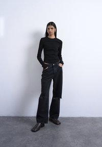 Ženska z dolgimi temnimi lasmi nosi črno majico z dolgimi rokavi, ohlapne črne raztrgane hlače in rjave čevlje, stoji z rokami v žepih ob beli steni.