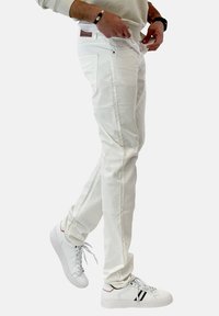 Bílé bavlněné džíny mají rovný střih, boční kapsy a malou hnědou koženou nášivku na zadním pasu. Nosič má na sobě bílé tenisky.