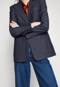 Tummansininen bleiseri, jossa on teksturoidun kankaan pinta, oranssit ompeleet, suuret etutaskut ja napin yksityiskohdat. Yhdistetty sinisiin farkkuihin.