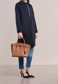Persona che indossa un cappotto blu navy, jeans blu e tacchi neri, mentre tiene una borsa di pelle marrone contro uno sfondo beige semplice.