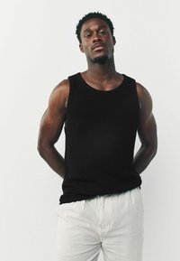 Zwarte gebreide tanktop met een ronde hals, textuurafwerking, gedragen met lichtgekleurde, relaxed-fit broeken. Het model poseert met de handen achter de rug.
