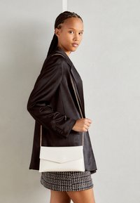 Vit crossbody väska med klaffdesign och kedjestropp, bärs ihop med en svart blazer och en mönstrad kjol med textur. Slätt material, minimalistisk stil.