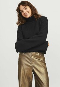 Maglione nero a collo alto in maglia con maniche lunghe, abbinato a pantaloni metallici dorati. Il maglione presenta una vestibilità rilassata e un tessuto testurizzato.
