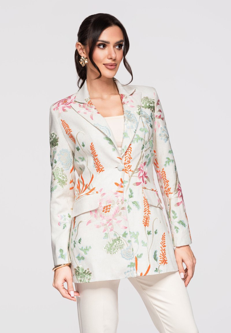 Mujer vestida con un blazer blanco con bordados florales multicolores y pantalones blancos a juego, de pie frente a un fondo liso y claro.
