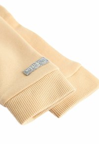 Beige gestrickte Pulloverärmel mit geripptem Muster und einem grauen Stoffetikett, das an einem Ärmel angenäht ist. Weiches, glattes Material in einheitlicher Farbe.