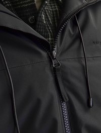 Chaqueta negra con un acabado mate y suave, que presenta una cremallera frontal, capucha y cordones ajustables. Se puede ver una tela de cuadros debajo.