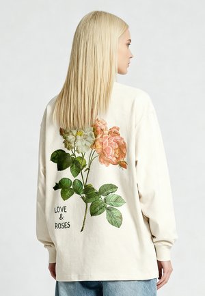 Vrouw met lang blond haar, die een creme kleurig shirt met lange mouwen draagt, voorzien van een grote bloemenprint en de tekst "LOVE & ROSES" op de rug.