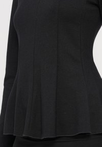 Schwarzes Langarmtop mit taillierter Passform und Peplum-Saum. Der Strickstoff hat eine glatte Textur mit sichtbaren Nahtdetails.