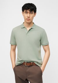 Νεαρός άνδρας με κοντά μαύρα μαλλιά που φοράει ένα πουκάμισο πόλο σε χρώμα φίκου και καφέ παντελόνι με καφέ ζώνη, στέκεται με τα χέρια στις τσέπες.