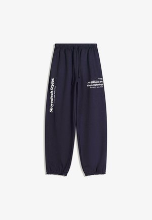 Laivastonsiniset collegehousut, joissa on joustava vyötärö ja resorit lahkeissa, valkoinen teksti vasemmassa reidessä ja oikeassa lahkeessa, jossa lukee "Shoreditech Styles" sekä inspiroivia lauseita.