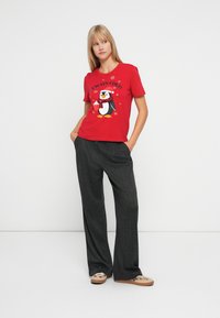 Ung kvinde iført en rød kortærmet T-shirt med en pingvin og teksten "Altid Kold", grå løse bukser og beige sneakers, hænderne i lommerne.
