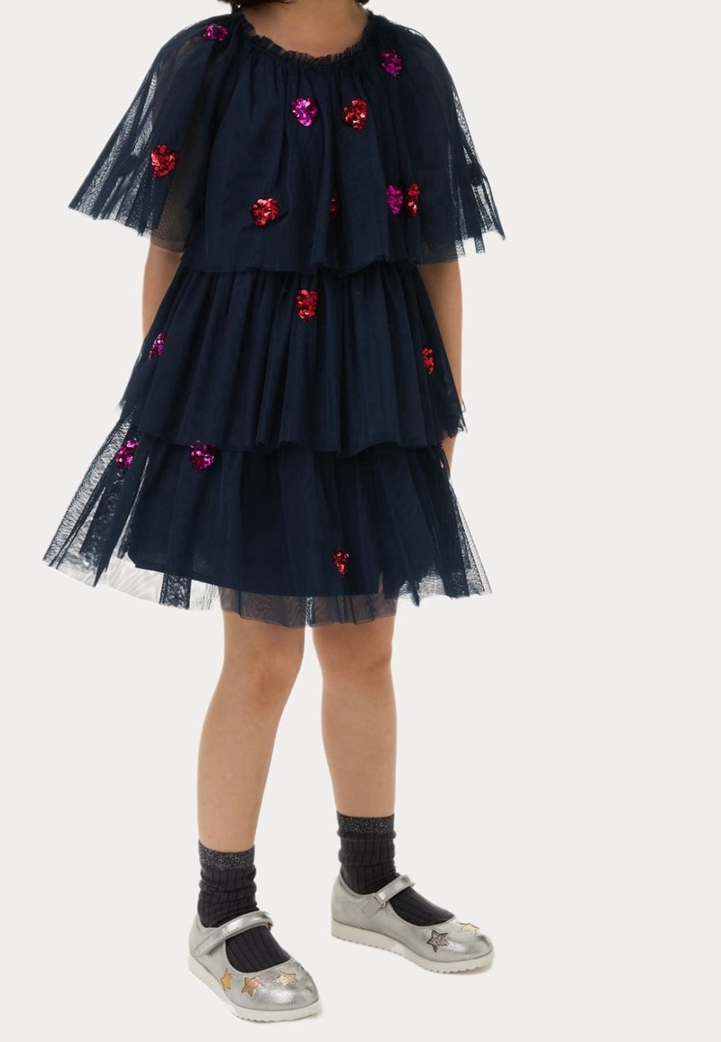Enfant portant une robe en tulle superposé bleu marine ornée de coeurs pailletés roses et rouges, chaussettes noires et chaussures argentées avec des décorations en forme d'étoiles.