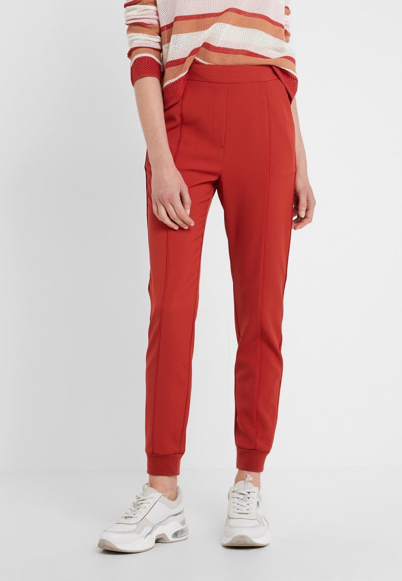 Bas du corps d'une personne portant un pantalon fuselé taille haute couleur rouille-orange, un top en maille rayé et des baskets blanches volumineuses sur fond blanc.